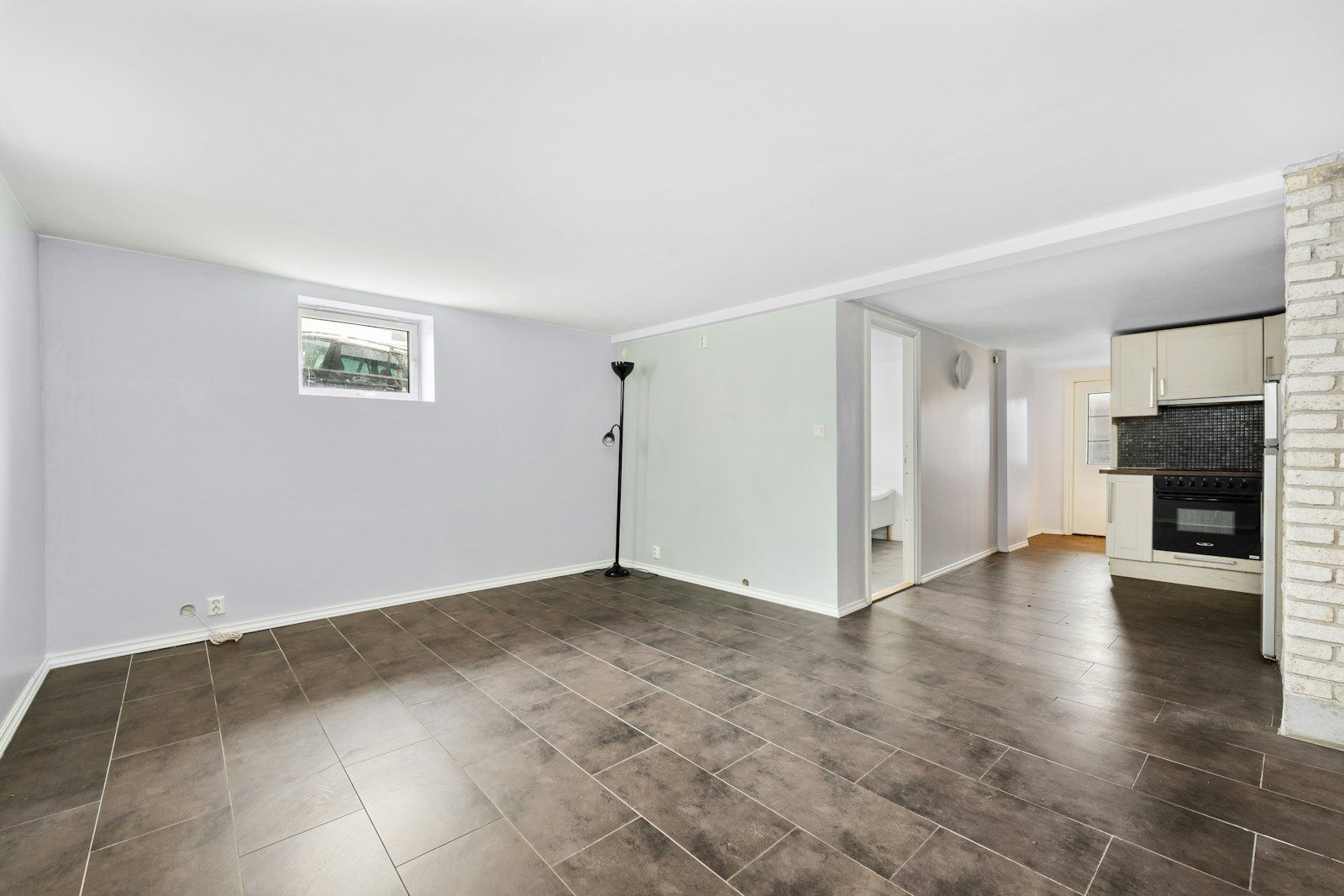 Empty living room with dark tiled floor, pale walls, a small high window and an open doorway to a kitchen